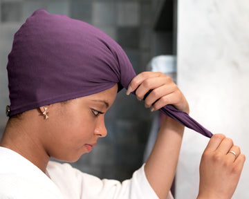 A woman wearing a Plum T-Shirt Hair Towel Hood for Curly, Wavy, and Straight Hair, adjusting it with both hands in a bathroom setting.