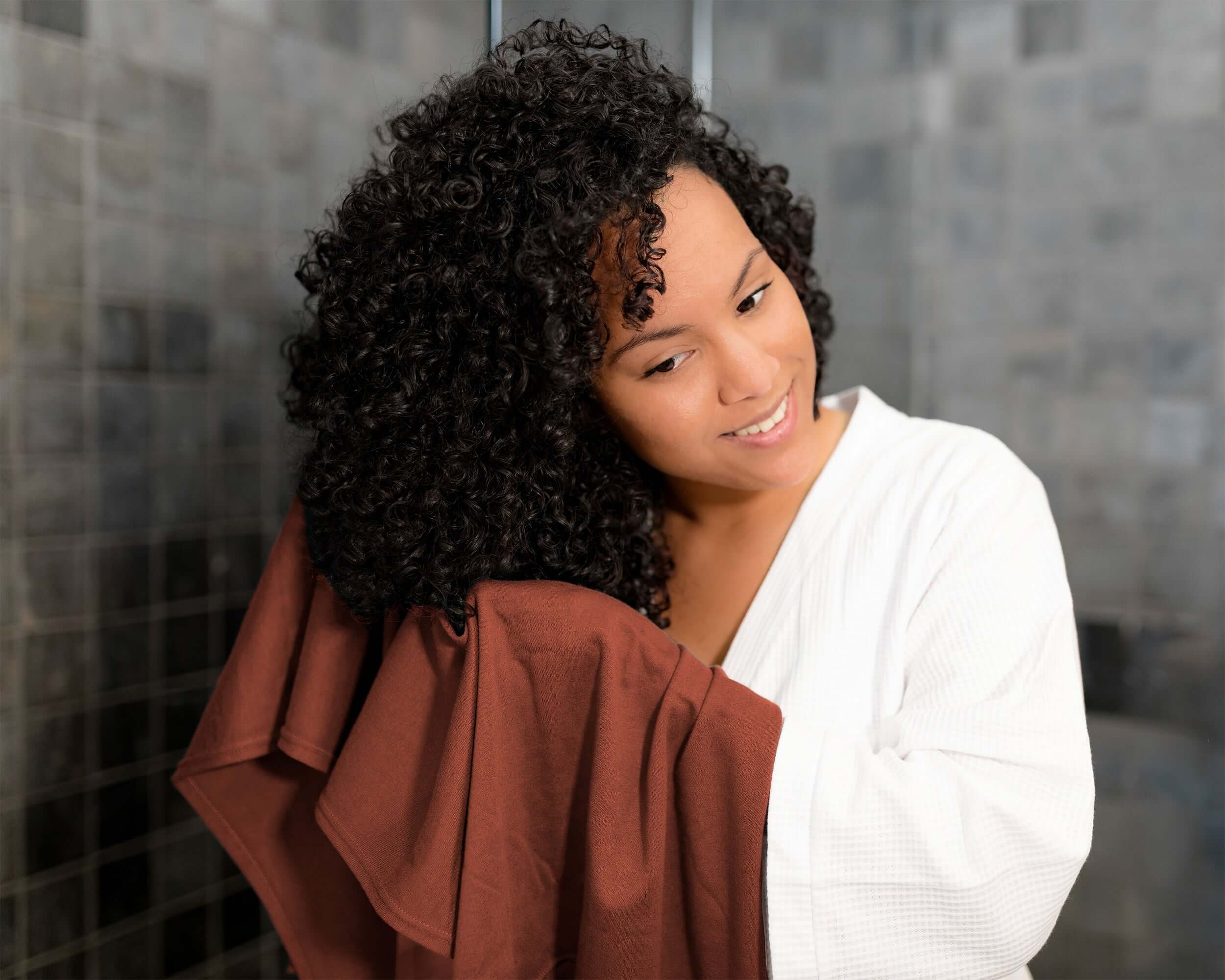 Woman with curly hair wrapped in a t-shirt hair towel against a tiled wall background