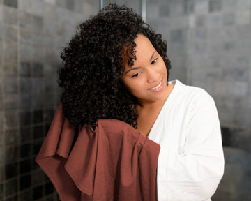 Woman with curly hair wrapped in a t-shirt hair towel against a tiled wall background