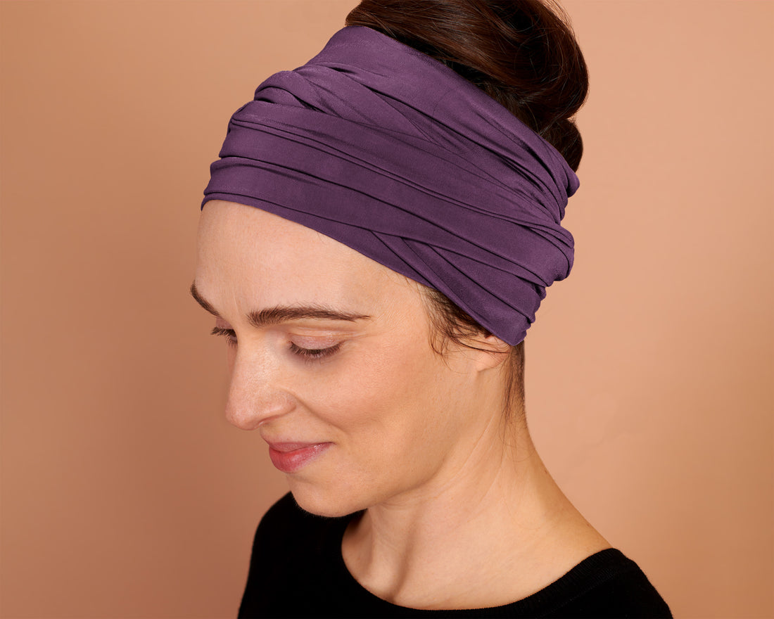 A woman wearing a Boho Head Wrap Wide Hair Wrap in light plum with her hair styled in a bun, shown in profile against a solid warm beige background.