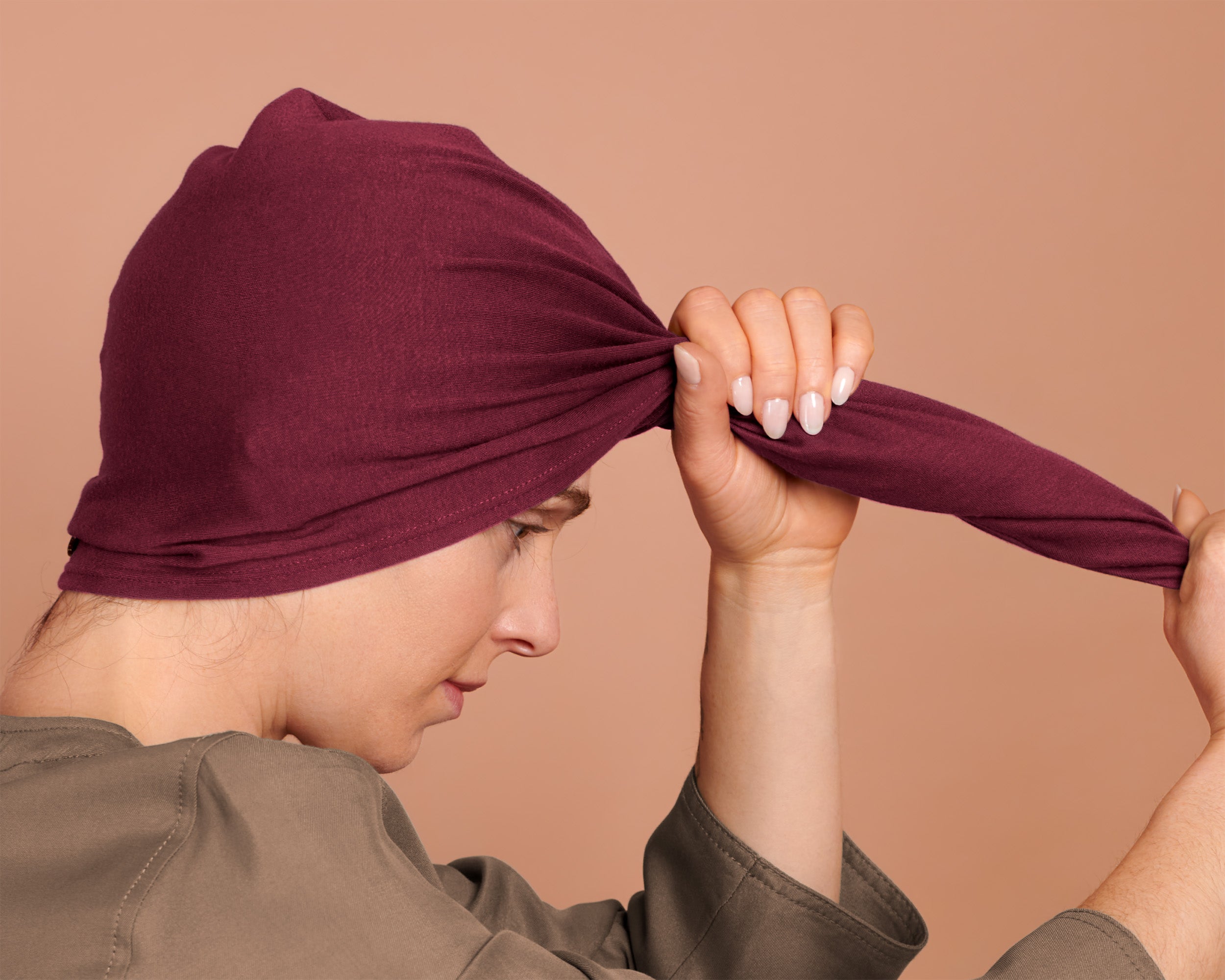 A woman  has wrapped the red burgundy t-shirt hair towel wrap hood around their head, displaying the towel in use, against a solid warm beige background.