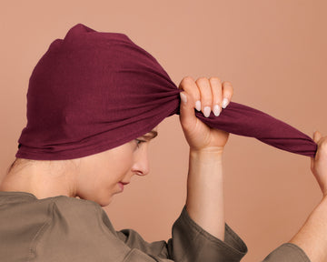 A woman  has wrapped the red burgundy t-shirt hair towel wrap hood around their head, displaying the towel in use, against a solid warm beige background.