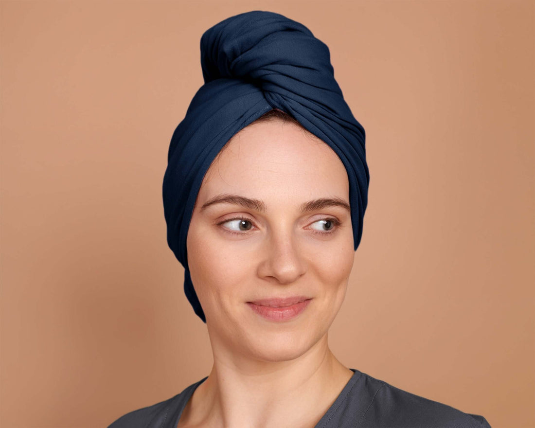 A person with curly hair wrapped in a marine blue t-shirt hair towel, standing against a beige background.