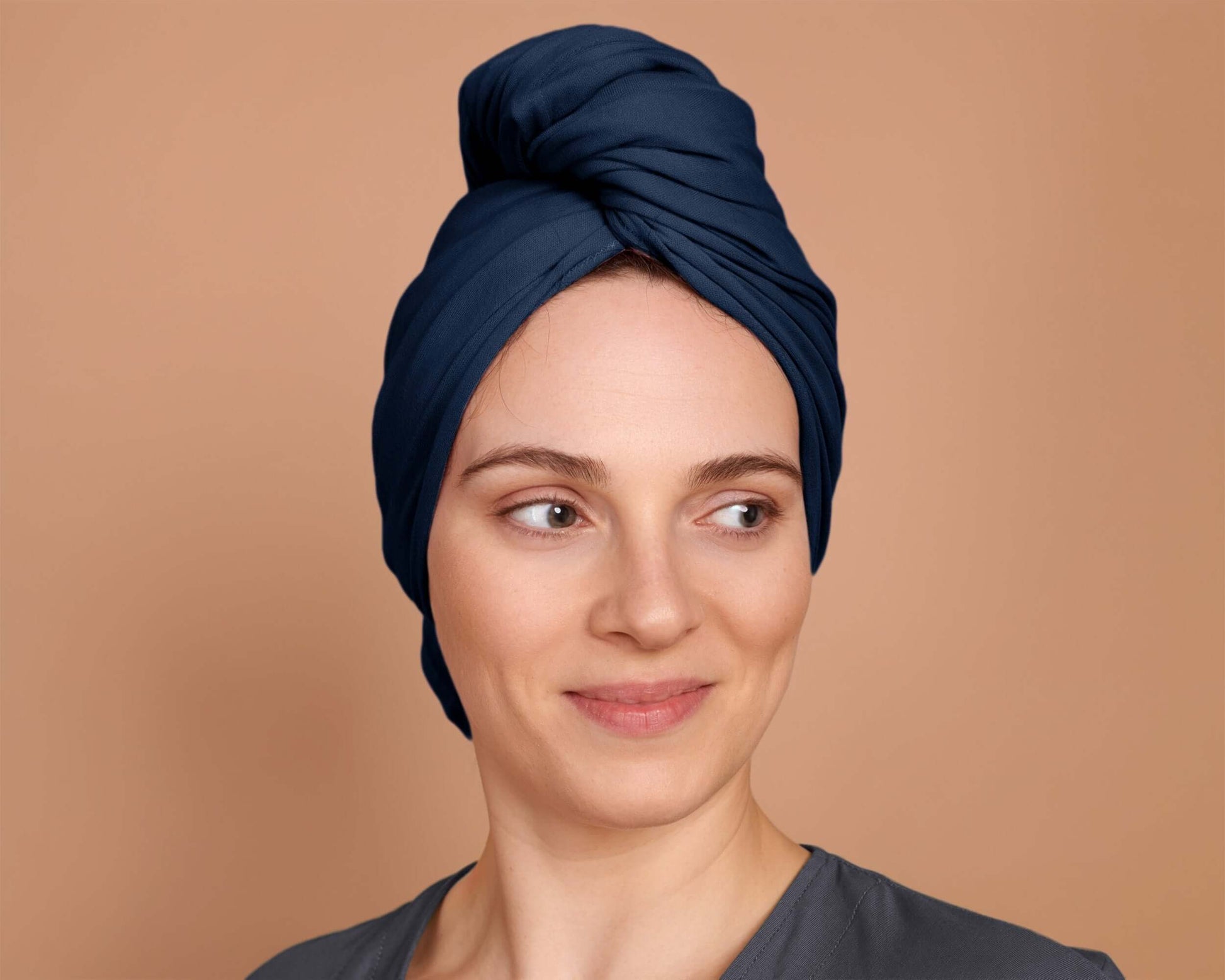 A person with curly hair wrapped in a marine blue t-shirt hair towel, standing against a beige background.