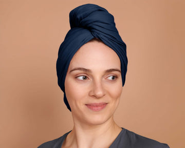 A person with curly hair wrapped in a marine blue t-shirt hair towel, standing against a beige background.
