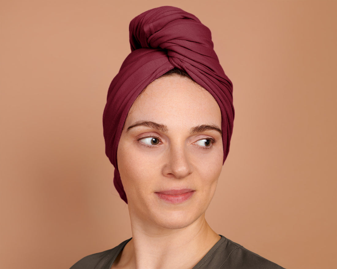 A woman  has wrapped the red burgundy t-shirt hair towel around their head, displaying the towel in use, against a solid warm beige background.