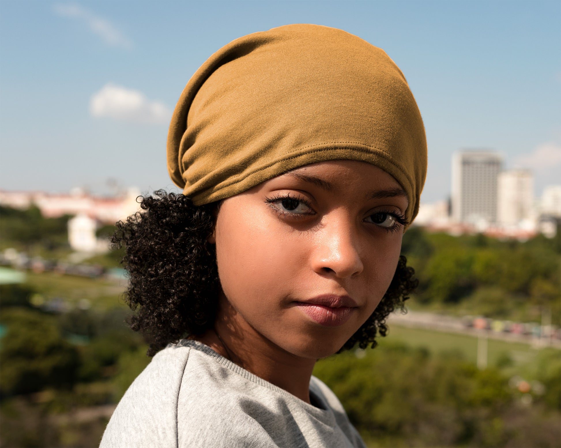 A close-up portrait of a young woman wearing a Satin Lined Beanie for Women and Men - Soft and Warm Beanie with Satin Lining to Protect Hair in yellow, with an outdoor city and park background.