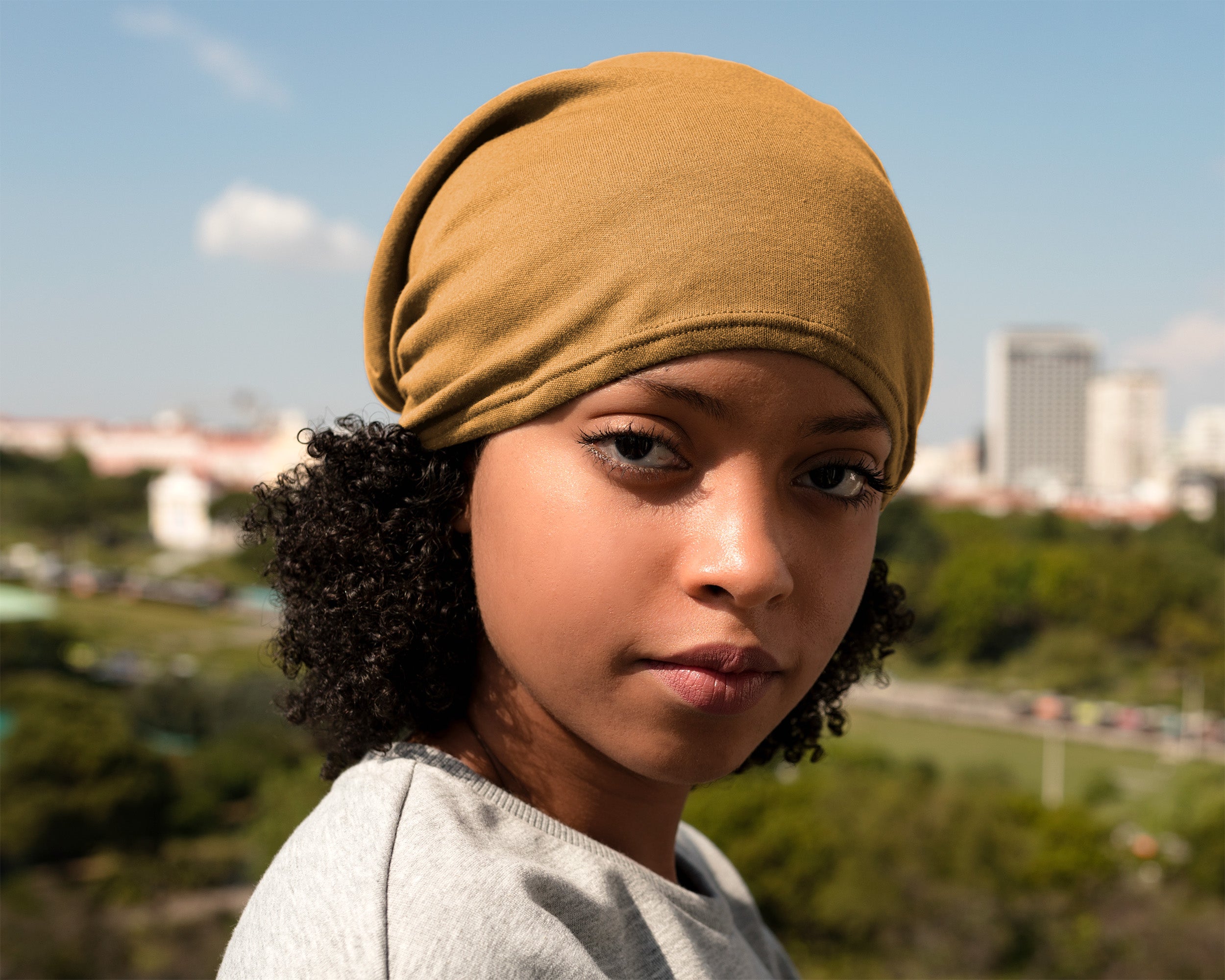 A close-up portrait of a young woman wearing a Satin Lined Beanie for Women and Men - Soft and Warm Beanie with Satin Lining to Protect Hair in yellow, with an outdoor city and park background.