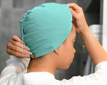 A woman wearing a T-shirt Eco-Friendly Hair Towel Hood, for Curly, Wavy, and Straight Hair - Soft, Absorbent in Aqua Blue, adjusting it with both hands. The cap has a small brown button closure on the back with a white drawstring.